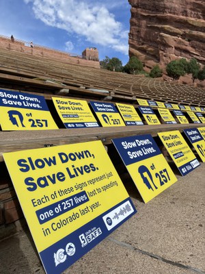 Yellow and blue lawn signs displayed in the seating area at Red Rocks Amphitheatre. Copy on the lawn signs reads "Slow down, save lives. Each of these signs represents just one of 257 lives lost to speeding in Colorado last year.” 
