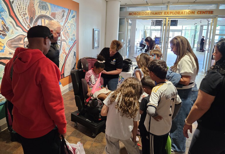 Children and adults gathered in a museum watching a woman properly secure a kid in a car seat. 