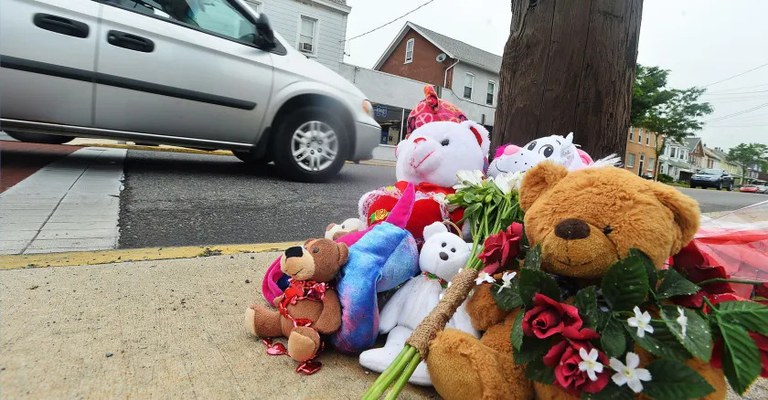 Stuffed animals and flowers at a roadside memorial for pedestrians killed by drivers with a grey van driving past.