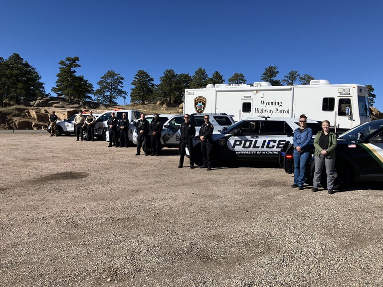 November LEL Photo Wyoming law enforcement officers and patrol cars lined up in a gravel parking lot with green trees and a blue sky.