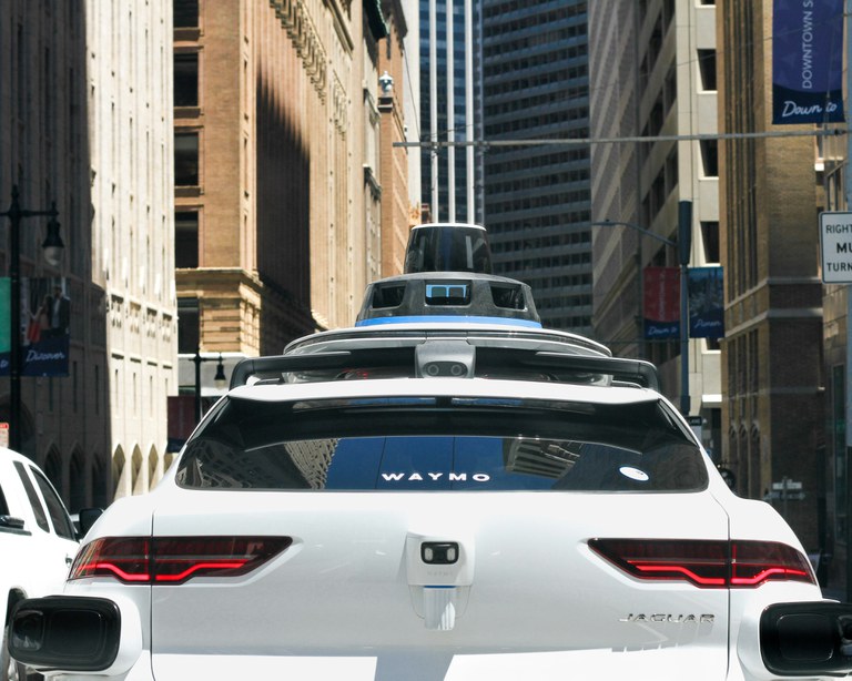Colorado requires drivers to be responsible White Waymo vehicle on an urban street with tall buildings.