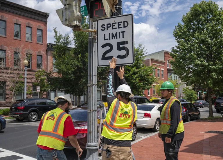 City workers in yellow vests installing a 25 mph sign near an urban street.