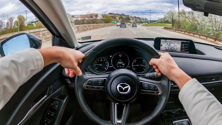 Safety benefits stack up from driver assistance features Interior view of a Mazda SUV from the driver’s seat with a person's hands on the steering wheel and cars on the roadway ahead.