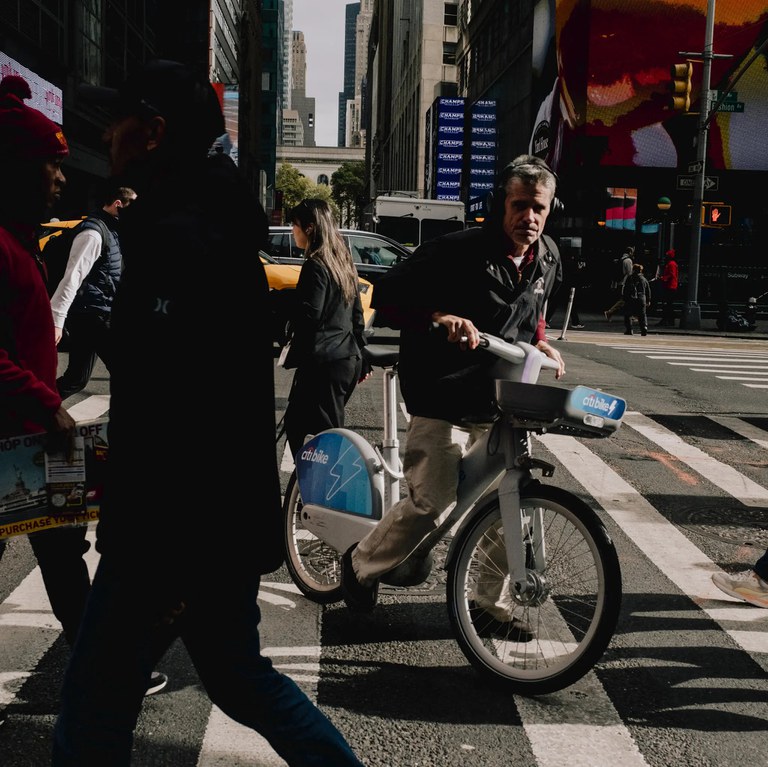 Bicyclist and pedestrians on a crowded city street with a yellow vehicle and tall buildings.