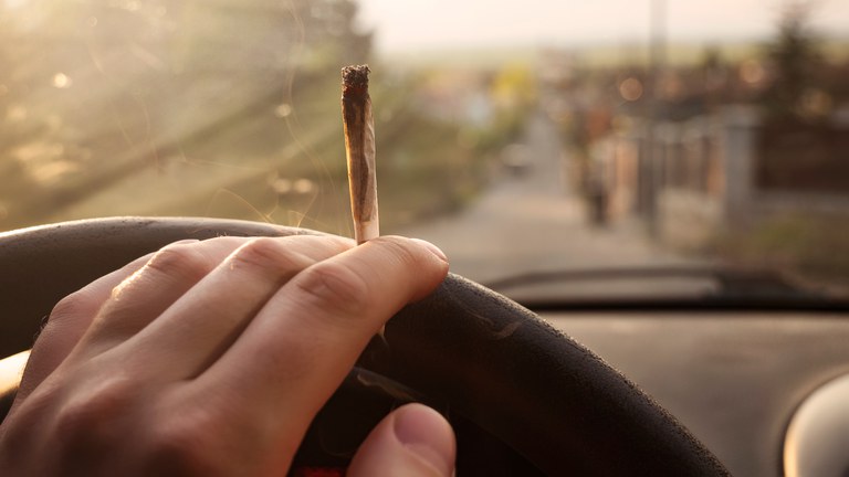 A lit joint of cannabis being held in hand, hand is also on steering wheel of a car. Outside of the window is a blurred road. 