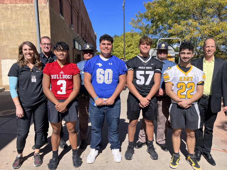 Nine people stand facing the camera for a group photo. Individuals who took part in a news conference on seat belt safety are pictured, including emergency responders in uniform, state law enforcement in uniform, Pueblo, Colorado student athletes in football jerseys, and a representative for the Colorado Department of Transportation.