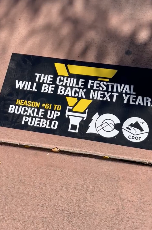 A vinyl sticker on a Pueblo sidewalk says “The Chile Fest will be back again next year. Reason number 61 to buckle up Pueblo.” Illustration of a seat belt and the Colorado Department of Transportation logo are visible behind the text.
