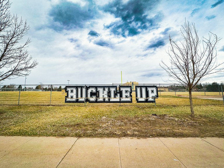 Safety first! “Buckle Up” message installed using cups in a chain-link fence at John F. Kennedy High School, Denver, CO