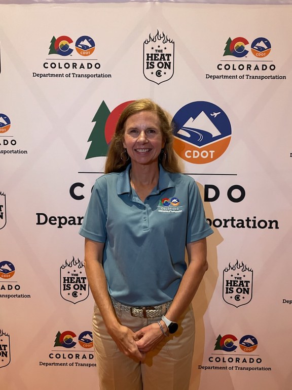 A woman, Cheryl Knibbe, standing in front of a backdrop that has The Heat Is On logos and a Colorado Department of Transportation logos