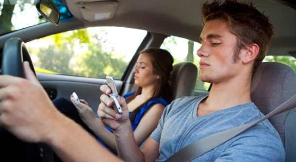 Man and woman in front seats of a car. Both are using cell phones, including the driver who has his other hand on the wheel. 